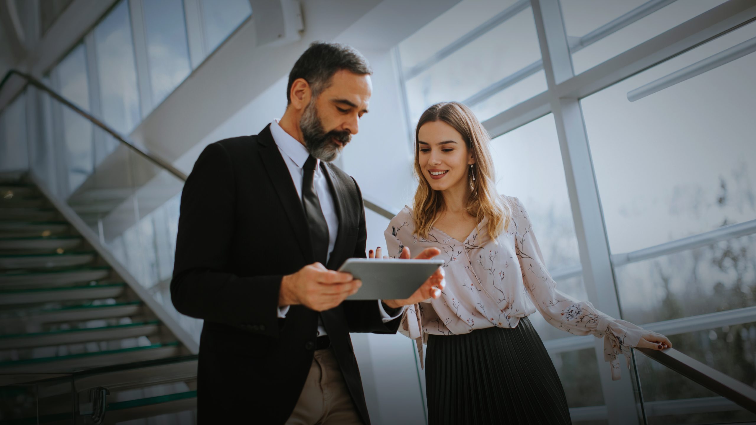 Two professionals dressed in formal attire are standing on a staircase inside a modern building with large windows. The man is holding and looking at a tablet. The woman is standing next to him.