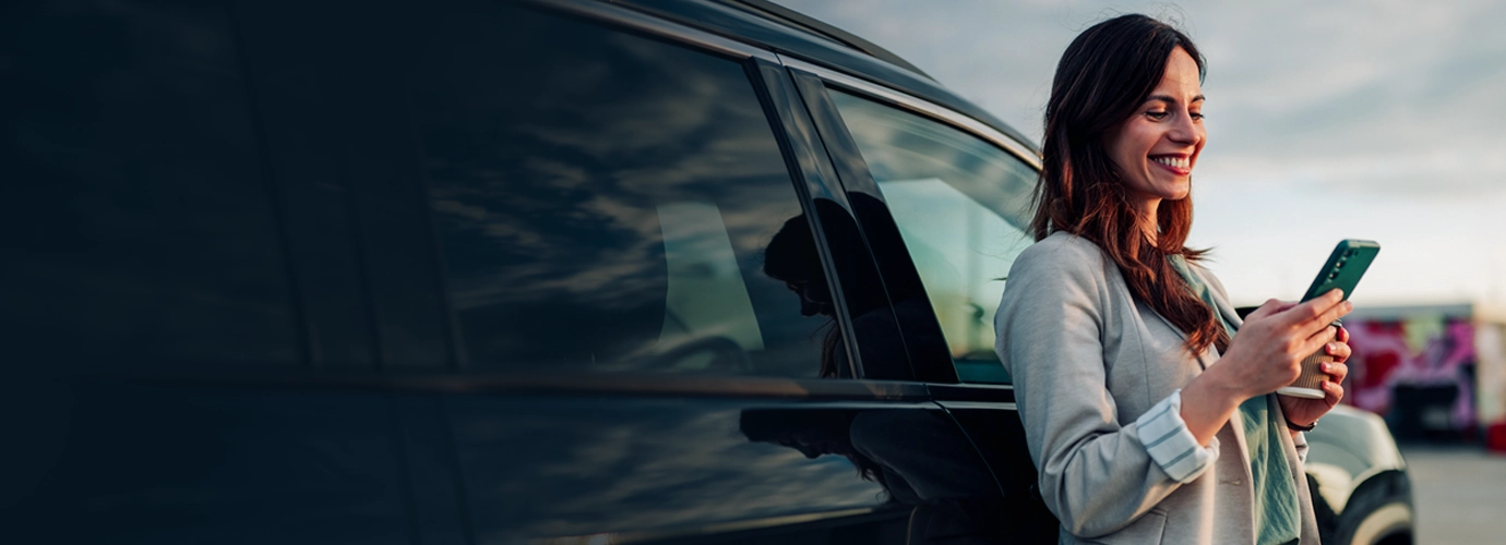 A woman stands beside a parked car, smiling as she uses her smartphone outdoors, suggesting a connected and convenient digital experience.